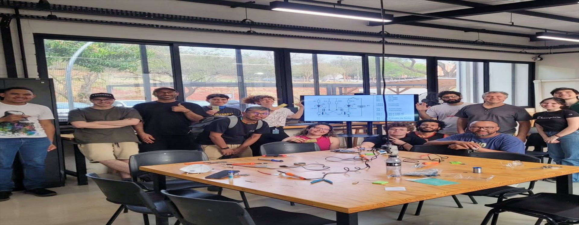 Grupo de participantes reunidos no Espaço Plasma durante oficina de solda eletrônica para iniciantes, com mesa de trabalho, ferramentas e componentes eletrônicos.
